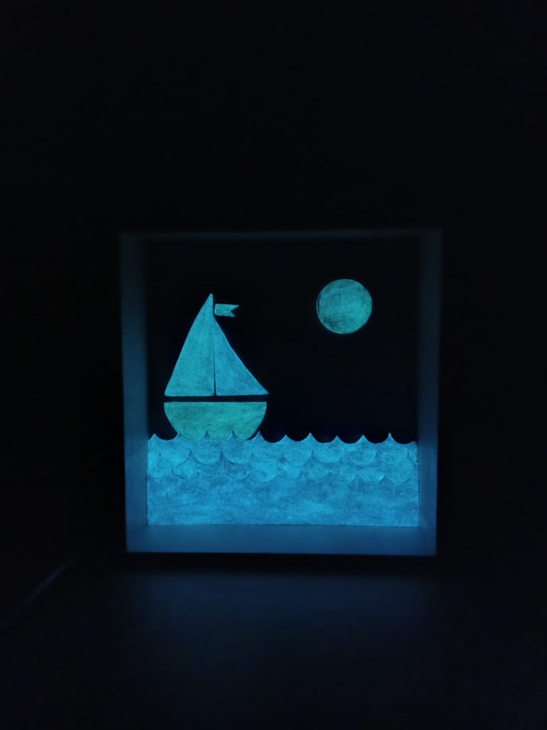 May include: A glow-in-the-dark wooden box with a sailboat and moon design. The sailboat is white with a yellow sail and is on a blue sea. The moon is yellow and is in the upper right corner of the box.
