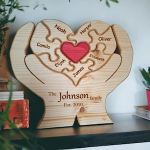Può includere: Puzzle di legno a forma di cuore con i nomi Noah, Camila, Elijah, James, Harper, Oliver, Elizabeth e un cuore rosso al centro. Il testo "The Johnson family Est. 2010." è inciso sotto il puzzle.