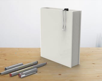 May include: A white blank notebook with a silver bookmark and four silver and gray pens on a wooden surface.