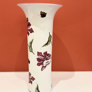 Puede incluir: Jarrón de cerámica blanca con borde rosa y forma acampanada. Está decorado con motivos florales burdeos y blancos, así como hojas verdes. Una pequeña mariposa está pintada cerca del borde. Un objeto decorativo para el hogar.