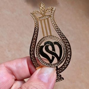 Puede incluir: Una horquilla dorada con forma de lira, con un diseño decorativo en forma de corazón en el centro. La horquilla tiene detalles intrincados y un acabado pulido, adecuada para varios peinados.