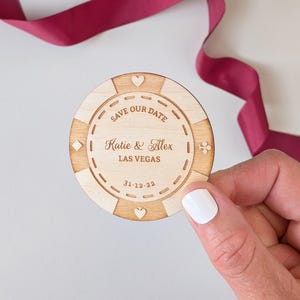 Peut inclure: Une annonce de mariage en bois en forme de jeton de casino. Le jeton porte l'inscription "Save Our Date", les noms "Katie & Alex" et "Las Vegas" avec la date "31.12.22". Le jeton est tenu près d'un ruban bordeaux.