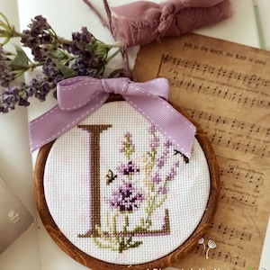 May include: A close-up of a hand-stitched embroidery hoop with a lavender floral design and the letter "L" in the center. The hoop is decorated with a purple ribbon and is resting on a piece of sheet music. The text "I Ricami della Natura" is visible at the bottom of the image.