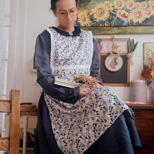 May include: A woman wearing a navy blue dress with a white floral print apron and white lace trim. She is holding a book and dried flowers in her lap. She is sitting on a wooden chair in front of a white wall with a painting of sunflowers and a framed chalkboard.
