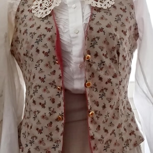 Può includere: Una blusa bianca con un colletto in pizzo all'uncinetto e un gilet a stampa floreale con bottoni dorati. Il gilet ha un bordo rosa.