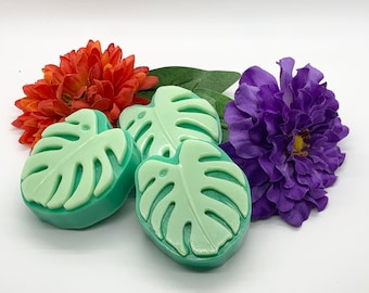 May include: Three green and light green leaf-shaped soaps. The soaps are arranged with orange and purple flowers and green leaves. The soaps have a tropical theme.