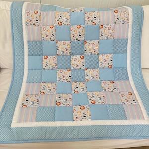 May include: A patchwork quilt featuring a repeating pattern of light blue squares, white squares with a colourful animal print, and striped squares in pastel tones. The quilt is bordered by a white trim and a light blue fabric with white polka dots.