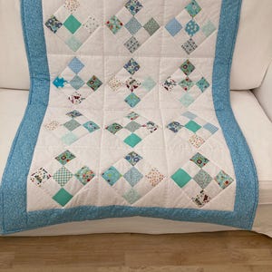 May include: A white baby quilt with a blue border and a diamond pattern made from various light blue and green fabrics with floral prints.