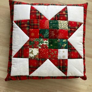 May include: A square throw cushion featuring a patchwork star design. The central star is composed of various red, green, and white fabric squares with holiday-themed patterns, including poinsettias, holly, and gold stars, surrounded by white triangles.