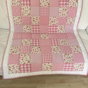 May include: A pink patchwork quilt with a white border. The quilt is made up of squares of different pink patterns, including gingham, stripes, and strawberry prints. The quilt is likely for a baby or child.