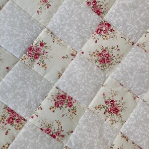 May include: Close-up of a quilted fabric with a repeating pattern. The design features alternating squares of floral bouquets in pink and cream, and grey squares with a white floral pattern. The quilt has a diagonal layout.
