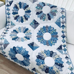 May include: A white quilt with a blue and white floral pattern. The quilt is made up of several squares, each with a different design. The quilt is folded and sitting on a wooden floor.