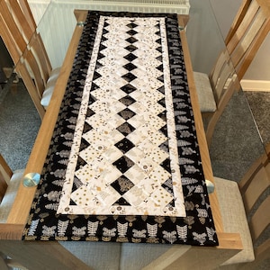May include: A quilted table runner featuring a central diamond pattern in black and white, framed by a black border with a gold leaf design. The runner is on a glass table.