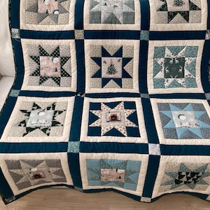 May include: A patchwork quilt featuring a grid of star-patterned blocks in shades of teal, navy blue, cream, and light blue. The fabric squares display various winter-themed prints including pine trees, deer, and fireplaces, separated by solid navy sashing.