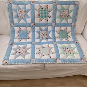 May include: A patchwork baby quilt with a star design. The quilt features light blue, white, and patterned squares with animal and geometric designs. The border is light blue with white polka dots and a striped edge.
