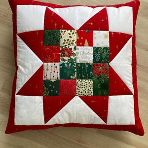 May include: A square quilted throw pillow featuring a central star pattern made of red and white fabric. The centre consists of a grid of sixteen small squares with holiday-themed prints including poinsettias, holly, and the text Merry Christmas.