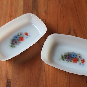 Peut inclure: Deux plats rectangulaires blanc cassé aux coins arrondis. Chaque plat présente un motif floral avec des fleurs bleues, rouges et violettes. Les plats sont posés sur une surface en bois.