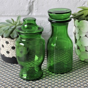 Peut inclure: Deux bocaux en verre vert avec couvercles et une bouteille en verre vert sont présentés sur une surface à motifs. Deux plantes succulentes en pot sont également présentes, l'une dans un pot à pois noirs et blancs et l'autre dans un pot blanc avec des accents circulaires.