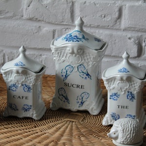 Puede incluir: Conjunto de tres botes de cerámica blanca con diseños florales azules y tapas. Los botes están etiquetados como "Cafe", "Sucre" y "The".