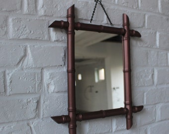 May include: A copper-colored mirror with a faux bamboo frame hangs on a white brick wall. The mirror is rectangular and has a simple design.