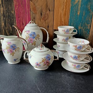 May include: A vintage tea set comprising a teapot, creamer, sugar bowl, and six teacups with saucers. The white porcelain set is adorned with blue and pink floral patterns and gold trim. The teapot is approximately 23 cm tall.