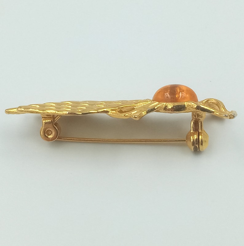 May include: A gold-toned brooch with an orange glass stone in the center. The brooch is shaped like a stylized leaf with a textured surface.