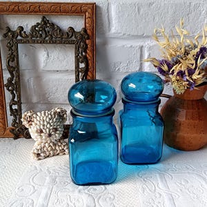 Peut inclure: Deux bocaux en verre bleu cobalt vintage avec couvercles arrondis. Les bocaux sont de forme carrée et reposent sur une surface blanche. Un cadre en bois décoratif et un petit ours en peluche en coquillages sont en arrière-plan.