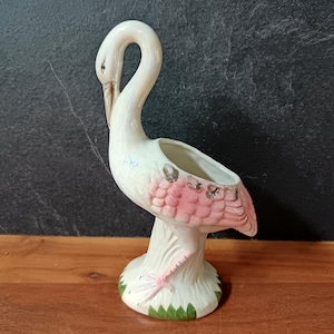Puede incluir: Una maceta de cerámica con forma de flamenco en blanco y rosa. El flamenco tiene un cuello largo y curvado y una pequeña abertura para plantar. Las alas están detalladas con plumas rosas y la base está decorada con hojas verdes.