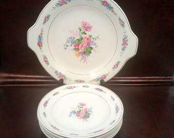 May include: A vintage white porcelain serving platter with handles and a stack of matching plates. Each piece features a floral bouquet in pink, blue, and yellow, with a gold rim. The platter has a decorative floral border.