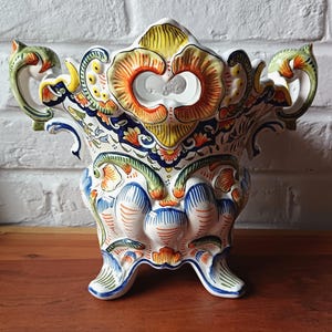 Op de afbeelding: Een handbeschilderde keramische vaas met een levendig bloemmotief. De vaas heeft een witte basis met kleurrijke accenten van geel, oranje, blauw en groen. Hij heeft twee handvatten en staat op drie poten, wat een gedetailleerde, sierlijke stijl laat zien.