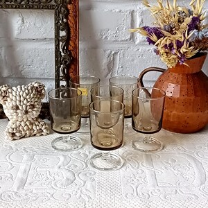 Könnte beinhalten: Eine Sammlung von Vintage-Gläsern mit bernsteinfarbenen Stielen. Die Gläser haben ein klassisches Design mit einer abgerundeten Schale und einem klaren Stiel. Eine kupferfarbene Kanne mit getrockneten Blumen befindet sich im Hintergrund. Ein dekorativer Rahmen und ein Muschelbär sind ebenfalls vorhanden.