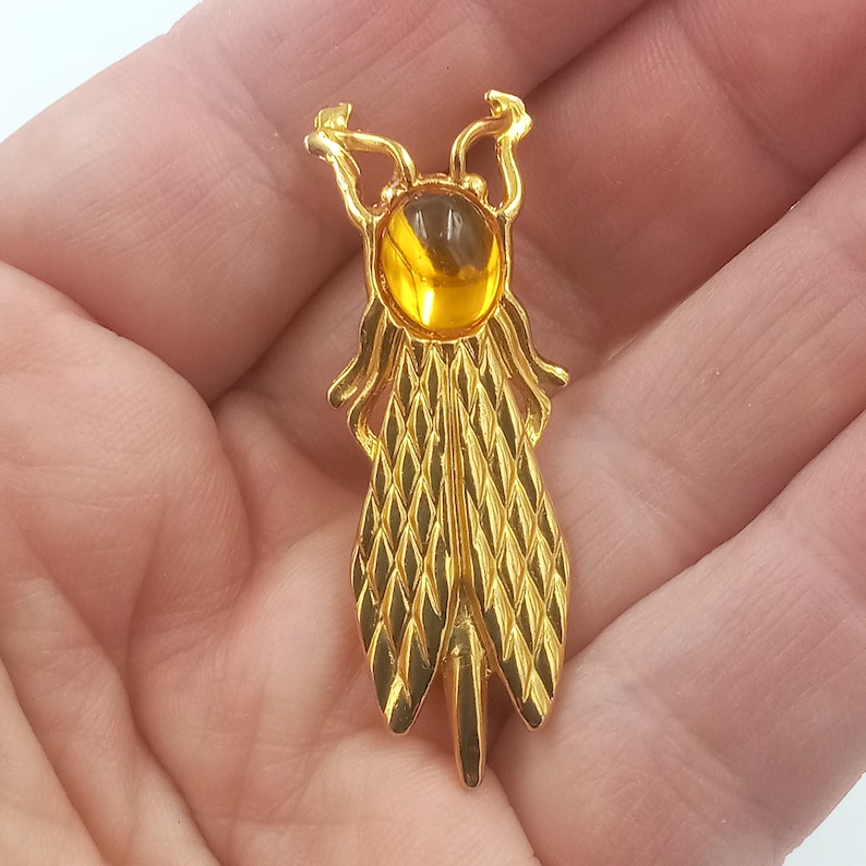 May include: A gold-toned brooch in the shape of a dragonfly with a yellow glass cabochon stone in the center.
