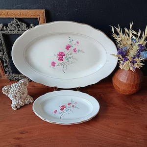 Puede incluir: Dos fuentes ovaladas blancas con bordes festoneados y ribete dorado. Cada fuente presenta un diseño floral con flores rosas y hojas verdes. Un pequeño jarrón marrón con flores secas está cerca.