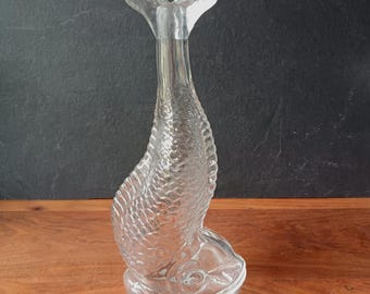 Puede incluir: Una botella de vidrio transparente con forma de pez, con cola acampanada y escamas texturizadas. La botella está sobre una superficie de madera con un fondo oscuro. La botella mide aproximadamente 30 cm de altura.