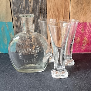 Peut inclure: Une carafe en verre transparent avec un motif de tournesol et trois verres à pied transparents. La carafe a un corps rond et un col court. Les verres ont une forme évasée et des bases décoratives. Les articles sont disposés sur une surface sombre.