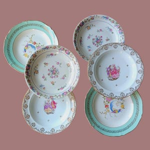 Può includere: Una collezione di sei piatti vintage in porcellana. I piatti presentano disegni floreali e di uccelli in vari colori, tra cui rosa, blu e giallo, con bordi dorati. Alcuni piatti hanno un bordo smerlato.