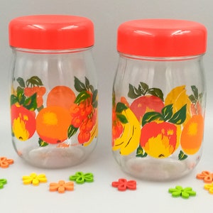 May include: Two glass jars with red lids and fruit designs. The jars have a clear glass body with a colourful design of fruits, including apples, oranges, bananas, and cherries. The jars are on a white surface with small colourful flower-shaped decorations.