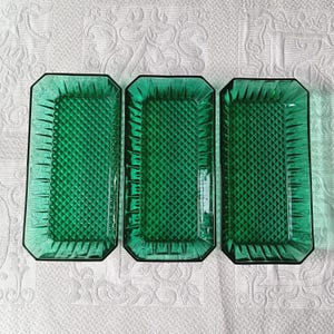Puede incluir: Tres bandejas rectangulares de vidrio verde esmeralda con un patrón de diamantes y borde negro. Las bandejas están dispuestas una al lado de la otra sobre una superficie blanca estampada. Las bandejas están vacías.