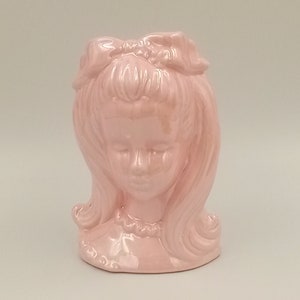 Peut inclure: Un vase en céramique rose en forme de tête de fille avec un nœud dans les cheveux.