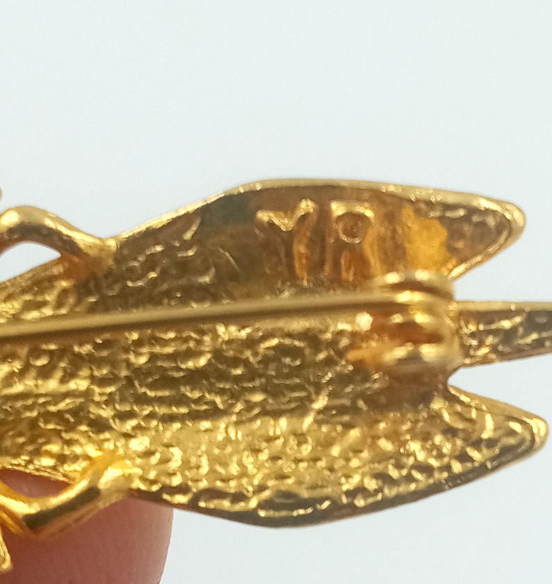 May include: Gold-toned metal dragonfly brooch with a textured surface and the inscription "VRI" on the back.