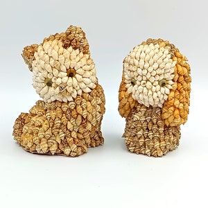 Puede incluir: Dos pequeñas figuritas de animales decorativas hechas de conchas marinas. Las figuritas son un gato y un perro, ambos con conchas blancas y marrones.