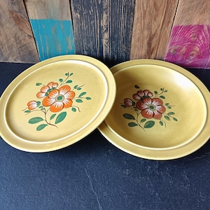 Peut inclure: Deux assiettes ovales jaunes avec des motifs floraux. Chaque assiette présente une fleur centrale avec des pétales orange et blancs, entourée de feuilles et de tiges vertes. Les assiettes ont un bord légèrement surélevé.