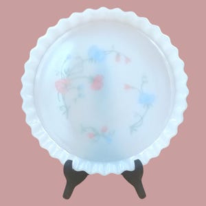Può includere: Un piatto da portata rotondo bianco con bordo smerlato. Il piatto presenta un motivo floreale rosa e blu. Il piatto è sostenuto da un supporto nero. Il piatto è probabilmente in vetro o ceramica.