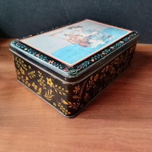 Può includere: Una scatola di latta nera rettangolare con coperchio incernierato. Il coperchio presenta un'immagine incorniciata di una donna con un vestito blu. I lati della scatola sono decorati con motivi floreali dorati. La scatola è su una superficie di legno.