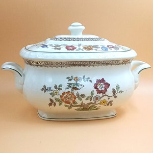 Może przedstawiać: Biała ceramiczna miska do serwowania z kwiatowym wzorem w odcieniach zieleni, brązu, czerwieni i żółci. Miska ma pokrywkę i dwa uchwyty.