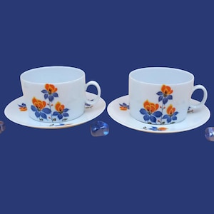 Puede incluir: Dos tazas de té y platillos blancos con un diseño floral en naranja y azul. Las tazas tienen forma redondeada con asa. Los platillos son redondos y tienen un patrón floral a juego. El fondo es azul liso.