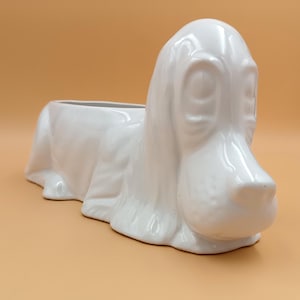 Könnte beinhalten: Weißer Keramik-Pflanztopf in Form eines liegenden Basset-Hundes. Der Topf hat eine rechteckige Öffnung zum Pflanzen.
