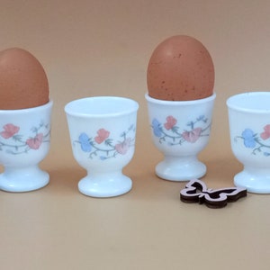 Può includere: Quattro portauova bianchi con disegni floreali, ognuno contenente un uovo marrone. I portauova hanno una base a piedistallo e sono decorati con fiori rosa e blu. Sono presenti anche due decorazioni a farfalla in legno.