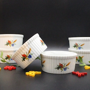 Op de afbeelding: Een set van zes witte keramische ramekins met verticale geribbelde zijkanten. Elke ramekin is versierd met een bloemenontwerp met rode, blauwe en gele bloemen. De ramekins zijn geplaatst op een donkere ondergrond.