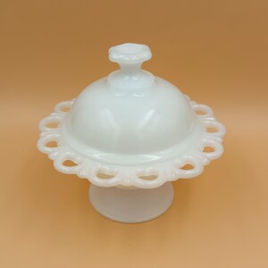 May include: A white, vintage-style covered dish with a scalloped edge and a pedestal base. The lid has a small, decorative handle. The dish is made of opaque glass and sits on a solid, round base. The overall design is elegant and classic.